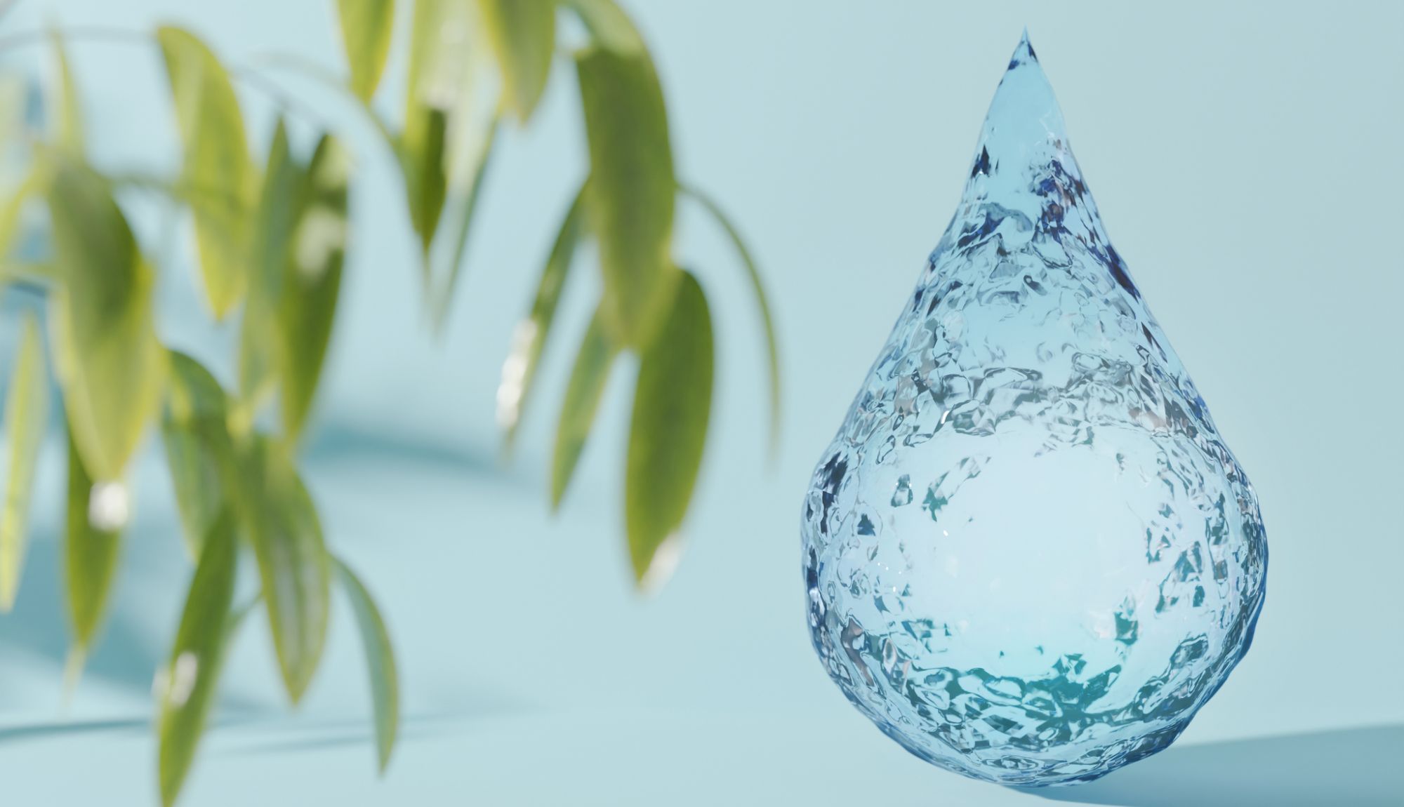 Nahaufnahme eines transparenten Wassertropfens mit strukturierter Oberfläche auf hellblauem Hintergrund, links im Bild unscharfe grüne Blätter.