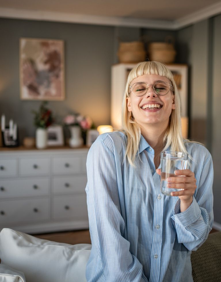 Junge Frau mit blonden Haaren und Brille sitzt lachend auf einem Sofa in einem gemütlich eingerichteten Wohnzimmer und hält ein Glas Wasser in der Hand.