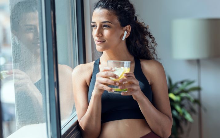 Junge Frau in Sportkleidung steht entspannt am Fenster, hält ein Glas Wasser mit Zitrone in der Hand und trägt kabellose Kopfhörer.